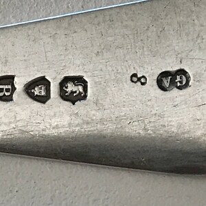 Może przedstawiać: Zbliżenie srebrnego naczynia z kilkoma cechami probierczymi. Oznaki obejmują tarczę z literą, tarczę z koroną, lwa, cyfrę 8 oraz litery GA. Naczynie ma odblaskową powierzchnię.