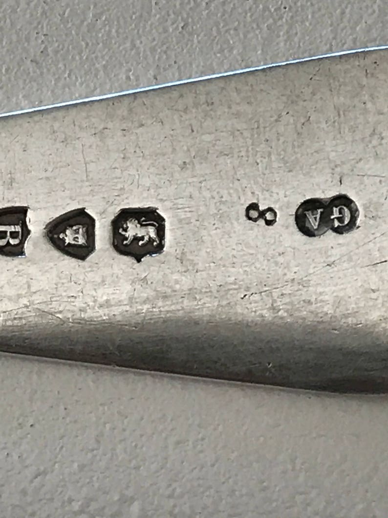 Może przedstawiać: Zbliżenie srebrnego naczynia z kilkoma cechami probierczymi. Oznaki obejmują tarczę z literą, tarczę z koroną, lwa, cyfrę 8 oraz litery GA. Naczynie ma odblaskową powierzchnię.