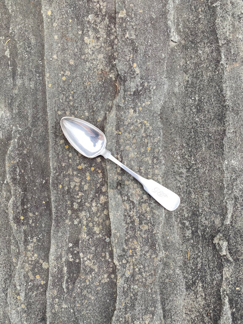 May include: A silver spoon with a rounded bowl and a long, slender handle. The spoon is lying on a textured, gray surface.