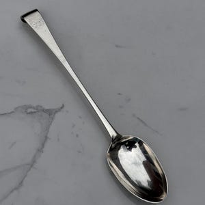 May include: A silver-toned metal spoon with a long, slender handle and a polished, oval-shaped bowl. The handle features a distinct, slightly curved end and a small, engraved rectangular detail near the top, showcasing a metallic reflective finish.