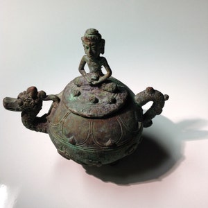 May include: A green bronze teapot with a dragon handle and a lid featuring a seated Buddha figure. The teapot has intricate details and a textured surface.
