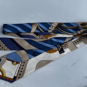 Puede incluir: Corbata de seda con un patr&oacute;n repetido de rayas azules y blancas, y dise&ntilde;os ecuestres beige y marr&oacute;n. La corbata est&aacute; doblada y tiene una peque&ntilde;a etiqueta negra con texto blanco.