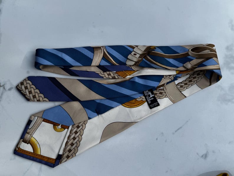 Puede incluir: Corbata de seda con un patr&oacute;n repetido de rayas azules y blancas, y dise&ntilde;os ecuestres beige y marr&oacute;n. La corbata est&aacute; doblada y tiene una peque&ntilde;a etiqueta negra con texto blanco.