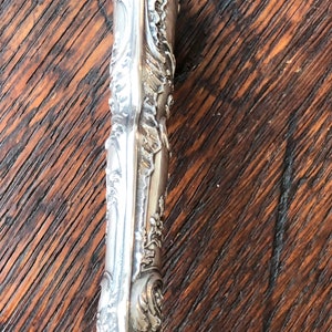 May include: Close-up of the ornate silver handle of a letter opener. The handle is highly detailed with intricate floral patterns and a smooth, polished finish.
