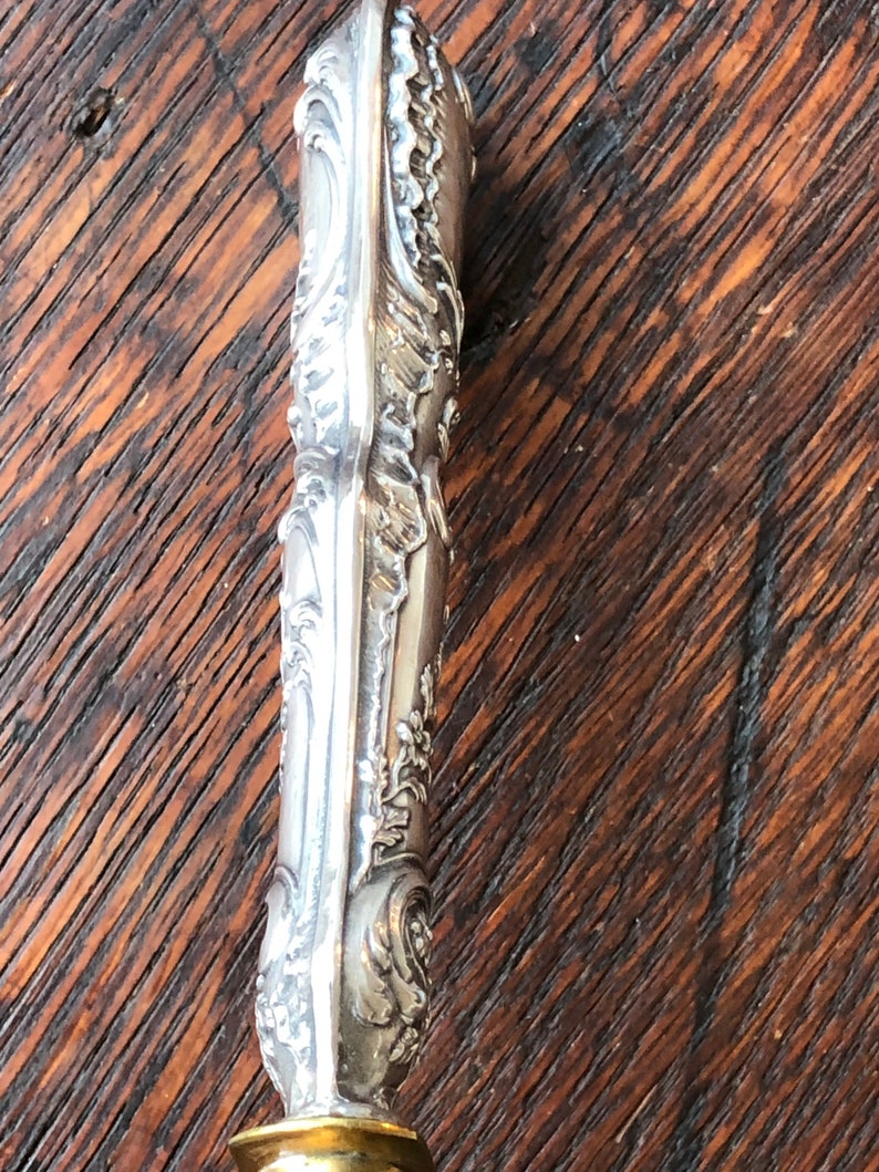 May include: Close-up of the ornate silver handle of a letter opener. The handle is highly detailed with intricate floral patterns and a smooth, polished finish.