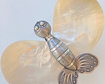Op de afbeelding: Een zilveren vlinder broche met een parelmoer vleugel ontwerp. De vlinder heeft een gedetailleerd lichaam met een gestreept patroon en een paar zwarte ogen. De tekst "DC SILVER" is zichtbaar op de vleugels.