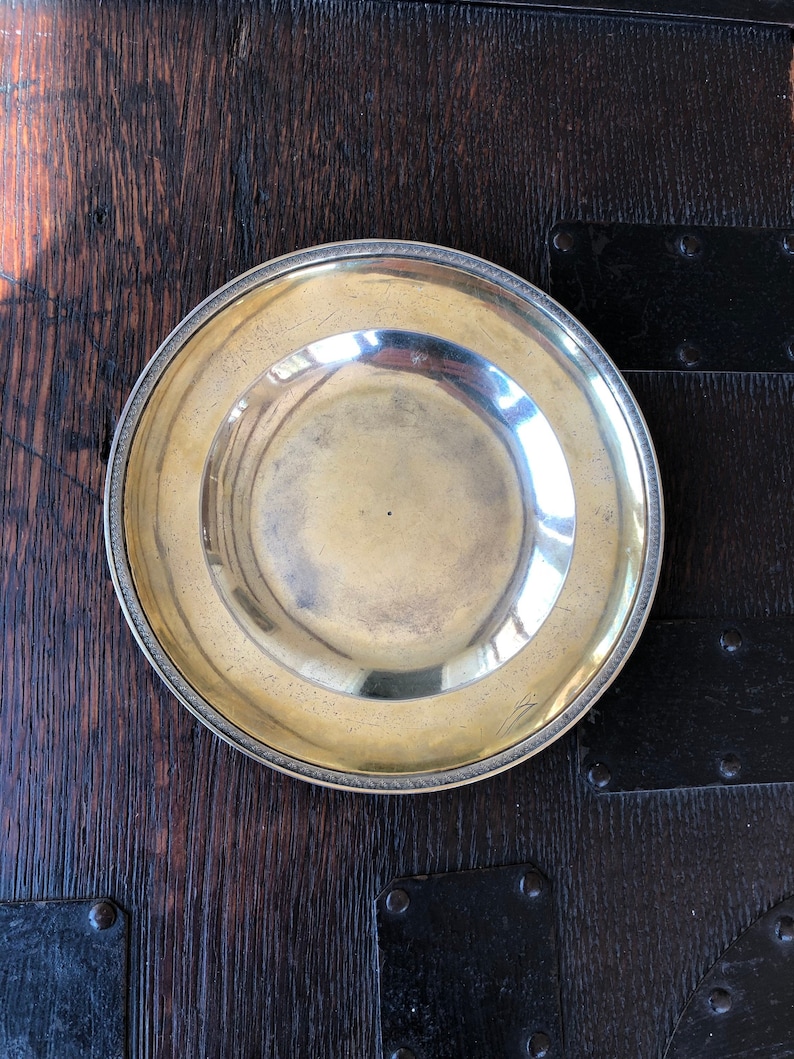 Puede incluir: Un plato redondo de color plateado con un borde elevado y una superficie ligeramente texturizada. El plato est&aacute; sobre una superficie de madera oscura.