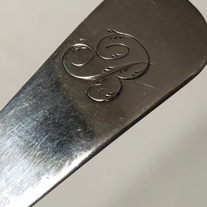 Puede incluir: Primer plano de una cuchara de plata con un dise&ntilde;o grabado en el mango. El dise&ntilde;o es una letra estilizada "S" con un adorno.