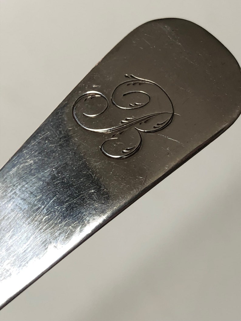 Puede incluir: Primer plano de una cuchara de plata con un dise&ntilde;o grabado en el mango. El dise&ntilde;o es una letra estilizada "S" con un adorno.