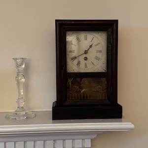 May include: An antique dark wood clock with a white face and Roman numerals. The clock is rectangular and sits on a dark base. A clear glass candlestick is to the left of the clock.