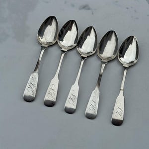 May include: Five antique silver spoons arranged on a light gray surface. Each spoon has an oval bowl and a slender handle with an engraved initial. The spoons reflect the light, highlighting their polished surfaces.