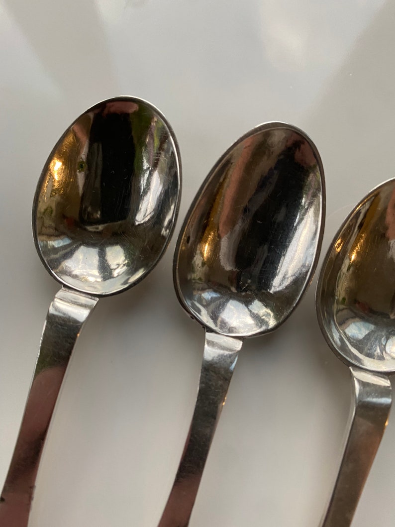 May include: Three silver spoons with rounded bowls and simple handles. The spoons are arranged in a row, with the bowls facing the viewer.