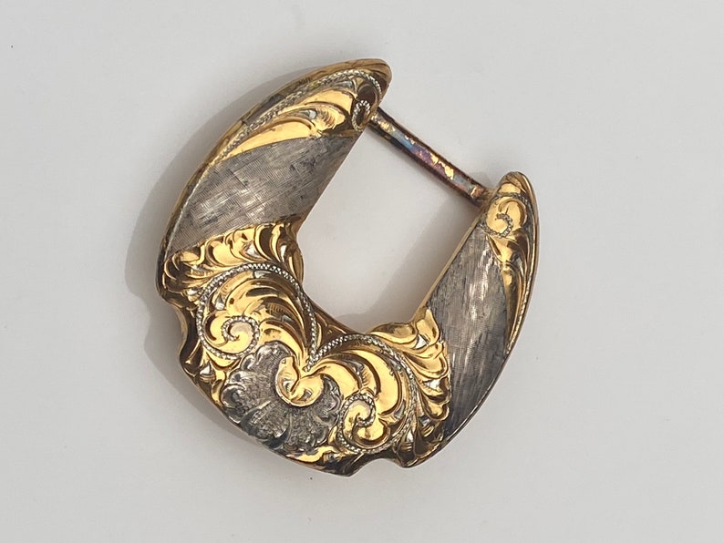 May include: A gold and silver belt buckle with intricate floral designs. The buckle is made of metal and has a polished finish.