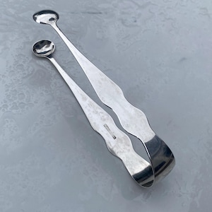 May include: Silver sugar tongs with a wavy design and rounded ends. The tongs have a hallmark on the side.