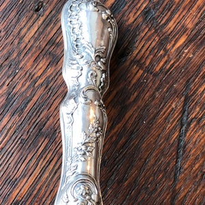 May include: Close-up of a silver handle with an ornate design. The handle is highly detailed with floral and scrollwork patterns.