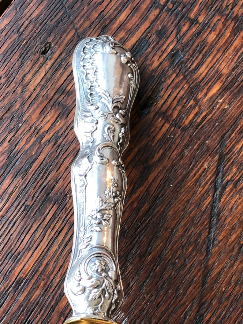 May include: Close-up of a silver handle with an ornate design. The handle is highly detailed with floral and scrollwork patterns.