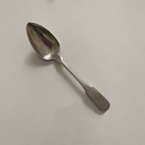 May include: A silver spoon with a rounded bowl and a simple, slightly curved handle. The handle is engraved with the initials "J.L.C."