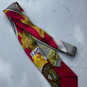 Puede incluir: Una corbata de seda roja con un patr&oacute;n detallado con detalles dorados y amarillos. La corbata tiene un borde gris y una etiqueta que dice "HERMES-PARIS". El dise&ntilde;o incluye elementos florales y en forma de cadena.