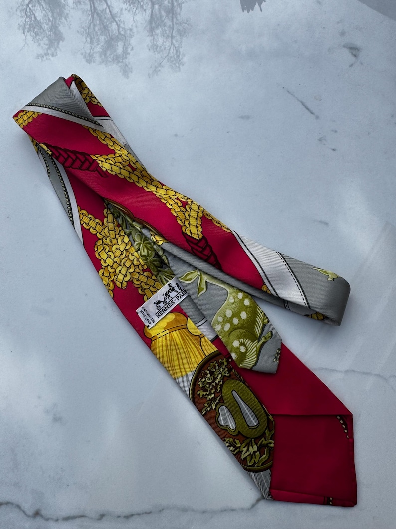 Puede incluir: Una corbata de seda roja con un patr&oacute;n detallado con detalles dorados y amarillos. La corbata tiene un borde gris y una etiqueta que dice "HERMES-PARIS". El dise&ntilde;o incluye elementos florales y en forma de cadena.