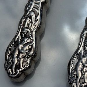 May include: Close-up of two ornate silver serving utensils. The handles feature detailed relief carvings of figures and floral patterns. The silver has a polished, reflective surface, and the design suggests a vintage or antique style.