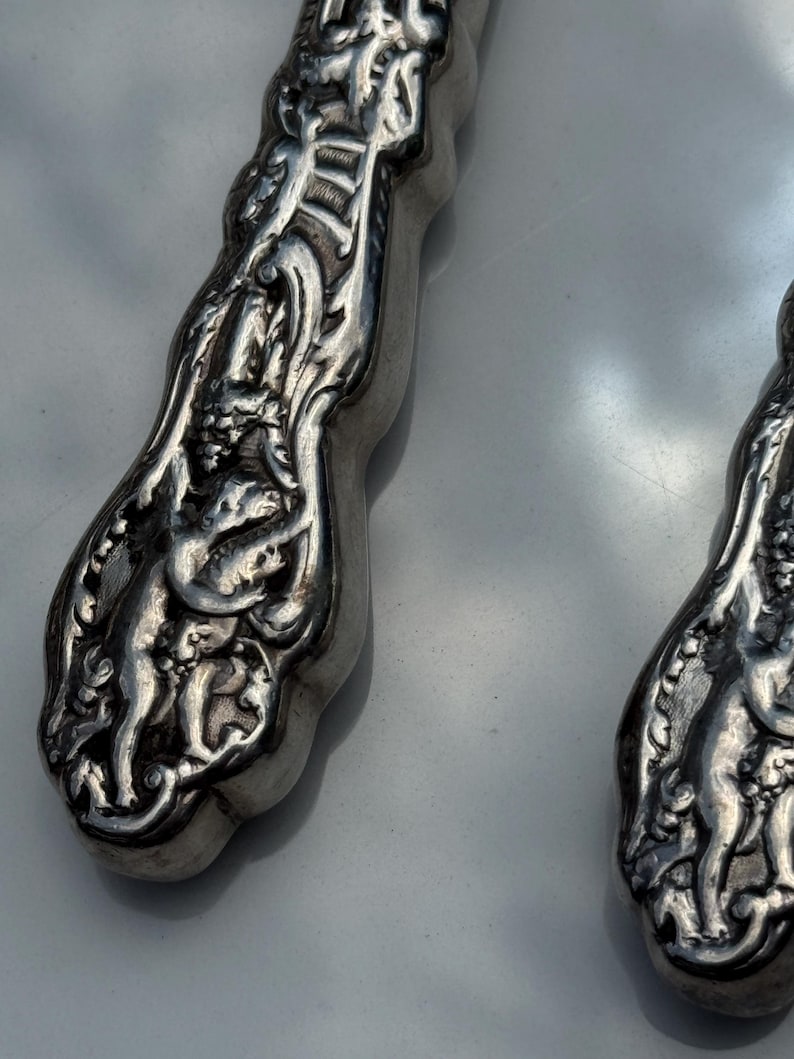 May include: Close-up of two ornate silver serving utensils. The handles feature detailed relief carvings of figures and floral patterns. The silver has a polished, reflective surface, and the design suggests a vintage or antique style.