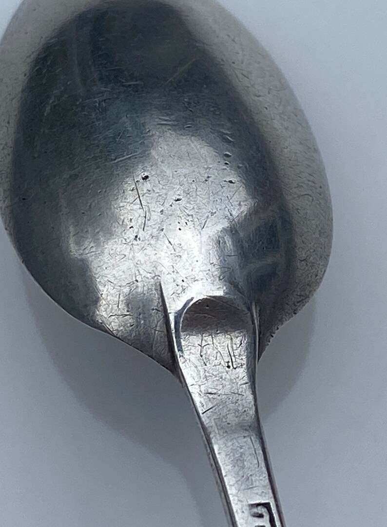 K&ouml;nnte beinhalten: Nahaufnahme der R&uuml;ckseite eines silbernen L&ouml;ffels mit abgenutzter Oberfl&auml;che und Markierungen. Der L&ouml;ffel hat eine abgerundete Schale und einen langen, schlanken Stiel.