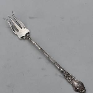 May include: A silver-toned metal cocktail fork featuring a three-pronged head with curved edges. The slender handle has a textured, bamboo-like midsection and terminates in an ornate, embossed floral pattern at the base of the handle.