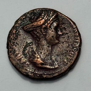 Puede incluir: Una moneda de bronce con el perfil de una persona con un tocado. La moneda tiene una superficie texturizada y una apariencia oscura y envejecida. El diseño es detallado, mostrando los rasgos faciales y los intrincados detalles del tocado.