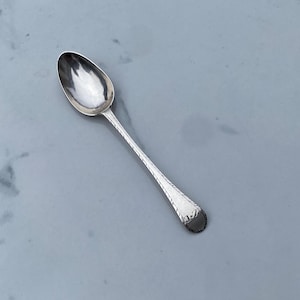 May include: A silver teaspoon with a rounded bowl and a simple, elegant design. The handle is decorated with a subtle pattern.