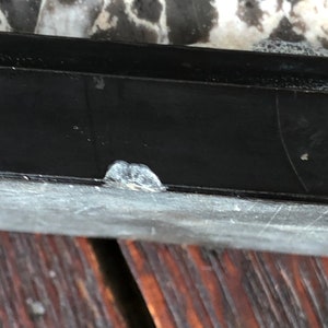 May include: Close-up of a black window frame with a small white paint chip on the bottom edge. The frame is above a dark brown wooden surface.