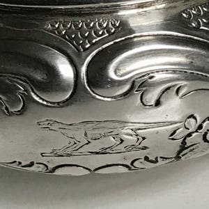 以下が含まれることがあります： 走る犬と花のモチーフが刻まれた銀製のボウルをクローズアップ。ボウルは滑らかで磨かれた表面をしています。