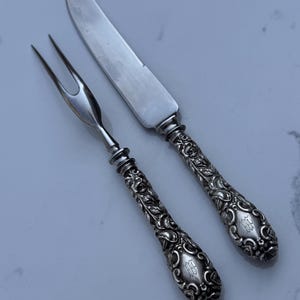 May include: A silver-toned serving fork and knife set featuring ornate, embossed floral and scrollwork patterns on the handles. Each handle includes a small, engraved monogram detail near the base, paired with smooth, polished metal blades.