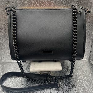 May include: A black rectangular handbag with a chain strap. The bag has a textured surface and a zipper closure. The chain strap is made of black metal links. The bag is displayed on a white platform against a silver background.