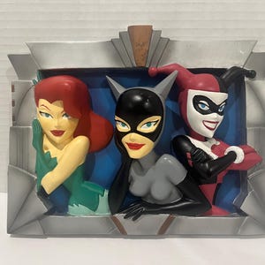 May include: A decorative art piece featuring three DC Comics characters: Poison Ivy, Catwoman, and Harley Quinn. The figures are in a stylized, cartoonish design, with vibrant colors and distinct features. The piece has a silver and gray art deco style frame.