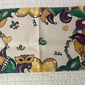May include: Rectangular off-white fabric with a Mardi Gras theme. The design includes masks, jester hats, and beads in green, purple, and gold. Decorative hearts and fleur-de-lis complete the festive look. The fabric is likely a placemat or decorative cloth.