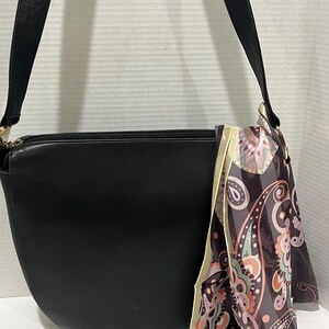 May include: Black leather shoulder bag with a curved design and a long strap. A decorative scarf with a paisley pattern in shades of pink, purple, and cream is draped over the bag.