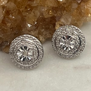 May include: Two round, silver-toned stud earrings. Each earring features a central, multi-faceted stone surrounded by a halo of smaller stones. The earrings are set against a neutral background.