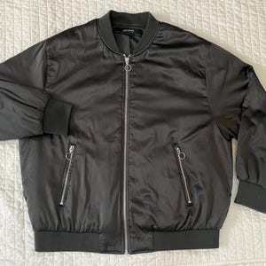 May include: Black bomber jacket with a zip-up front, two side pockets, and ribbed cuffs and hem. The jacket has a shiny, satin-like finish and a classic bomber silhouette. The jacket is a versatile outerwear piece.
