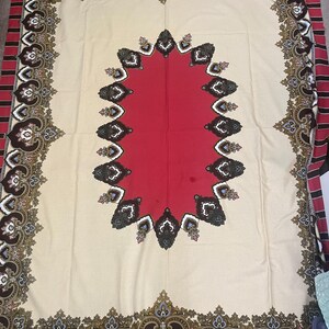 May include: A large, rectangular tablecloth with an ornate design. The center is red, surrounded by a cream-colored field. The border features a repeating pattern in brown, gold, and white, with a red and brown striped edge.