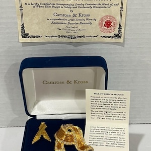 May include: A Camrose & Kross jewelry set featuring a gold-toned ribbon brooch and a smaller ribbon pin, presented in a blue velvet box. The set includes a certificate of authenticity. The brooch is adorned with clear crystals.