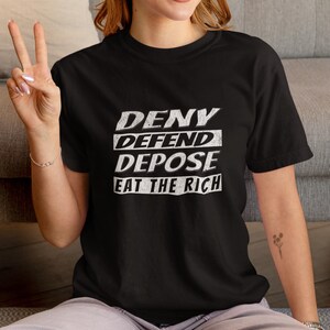 Può includere: Maglietta nera con testo bianco che recita "DENY DEFEND DEPOSE EAT THE RICH".