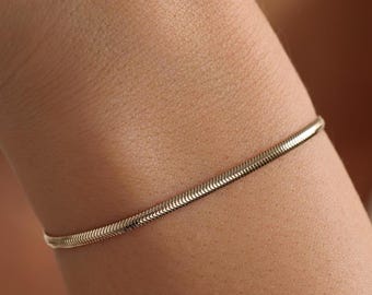 Può includere: Un bracciale a catena snake sottile color oro con un design a maglie intrecciate, lisce e flessibili. Il bracciale presenta una finitura metallica lucida che riflette la luce, creando un aspetto continuo, elegante e uniforme attorno al polso.
