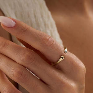 Puede incluir: Un anillo abierto de color dorado con las letras "S" y "E" grabadas en los extremos. El anillo se lleva en un dedo, y el fondo es de un color suave y neutro.