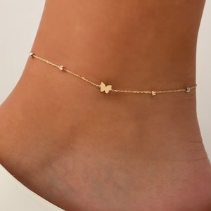 Pode incluir: Um tornozelo de ouro com um pingente de borboleta e pequenas contas. O tornozelo é delicado e feminino.