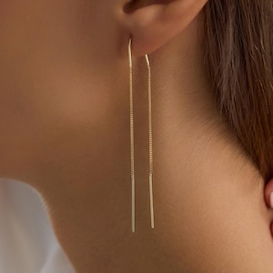 May include: A gold threader earring featuring a delicate chain that loops through the earlobe, ending in two slender, straight metal bars of equal length that hang vertically on either side of the earlobe, creating a minimalist and sleek appearance.
