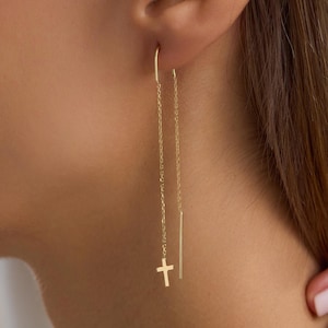 Könnte beinhalten: Ein goldener Durchziehohrring mit einer feinen Kette, die durch das Ohrläppchen verläuft. Ein Ende der Kette endet in einem kleinen, polierten goldenen Kreuzanhänger, während das andere Ende einen geraden, schlanken Goldstab für ein Design aufweist.