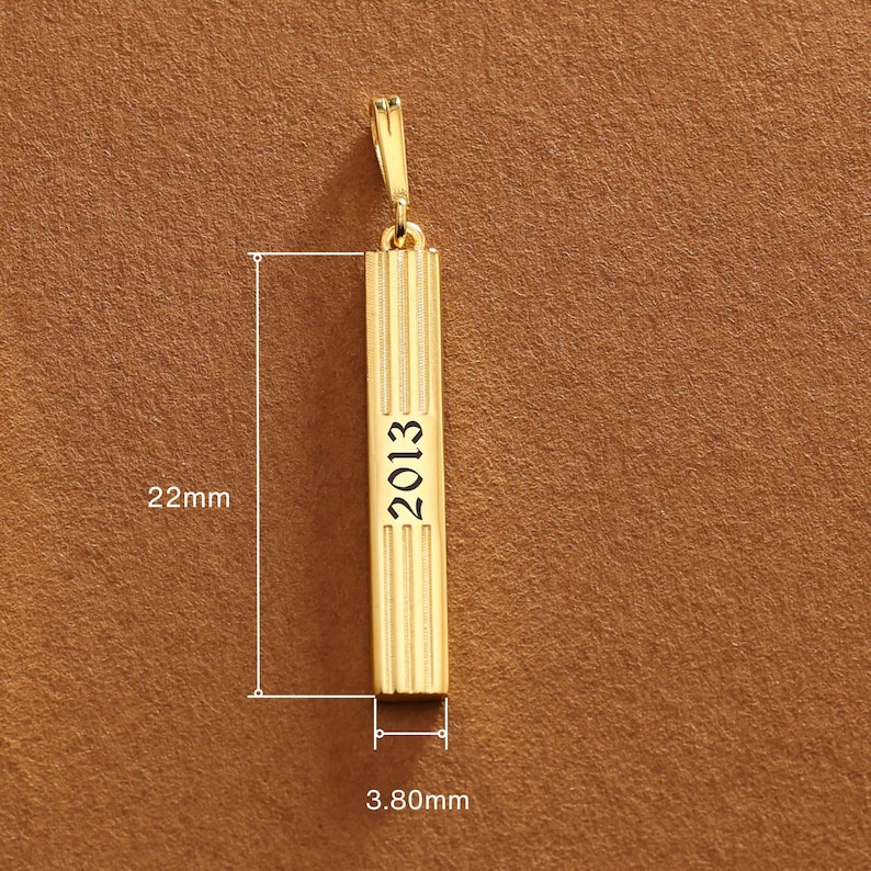 May include: A gold-colored rectangular pendant with vertical grooves and the year "2013" engraved on it. The pendant is 22mm long and 3.80mm wide. It has a small loop at the top for attaching to a chain.