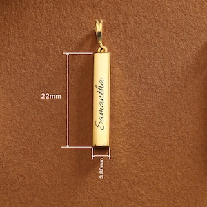 May include: A gold-colored rectangular pendant with the name "Samantha" engraved in a cursive font. The pendant is 22mm long and 3.80mm wide. It has a small loop at the top for attaching to a chain. The background is a solid brown color.