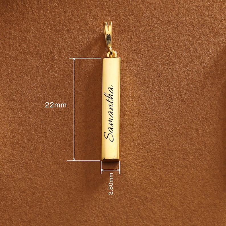 May include: A gold-colored rectangular pendant with the name "Samantha" engraved in a cursive font. The pendant is 22mm long and 3.80mm wide. It has a small loop at the top for attaching to a chain. The background is a solid brown color.