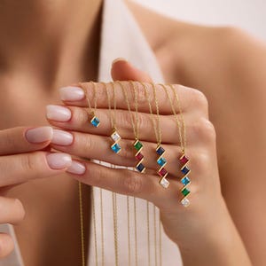 Puede incluir: Una colección de collares de oro con colgantes de piedras preciosas de colores. Cada collar presenta una fina cadena de oro y un colgante con piedras cuadradas de varios colores, como azul, verde, rojo y blanco. Las piedras están dispuestas en zigzag.